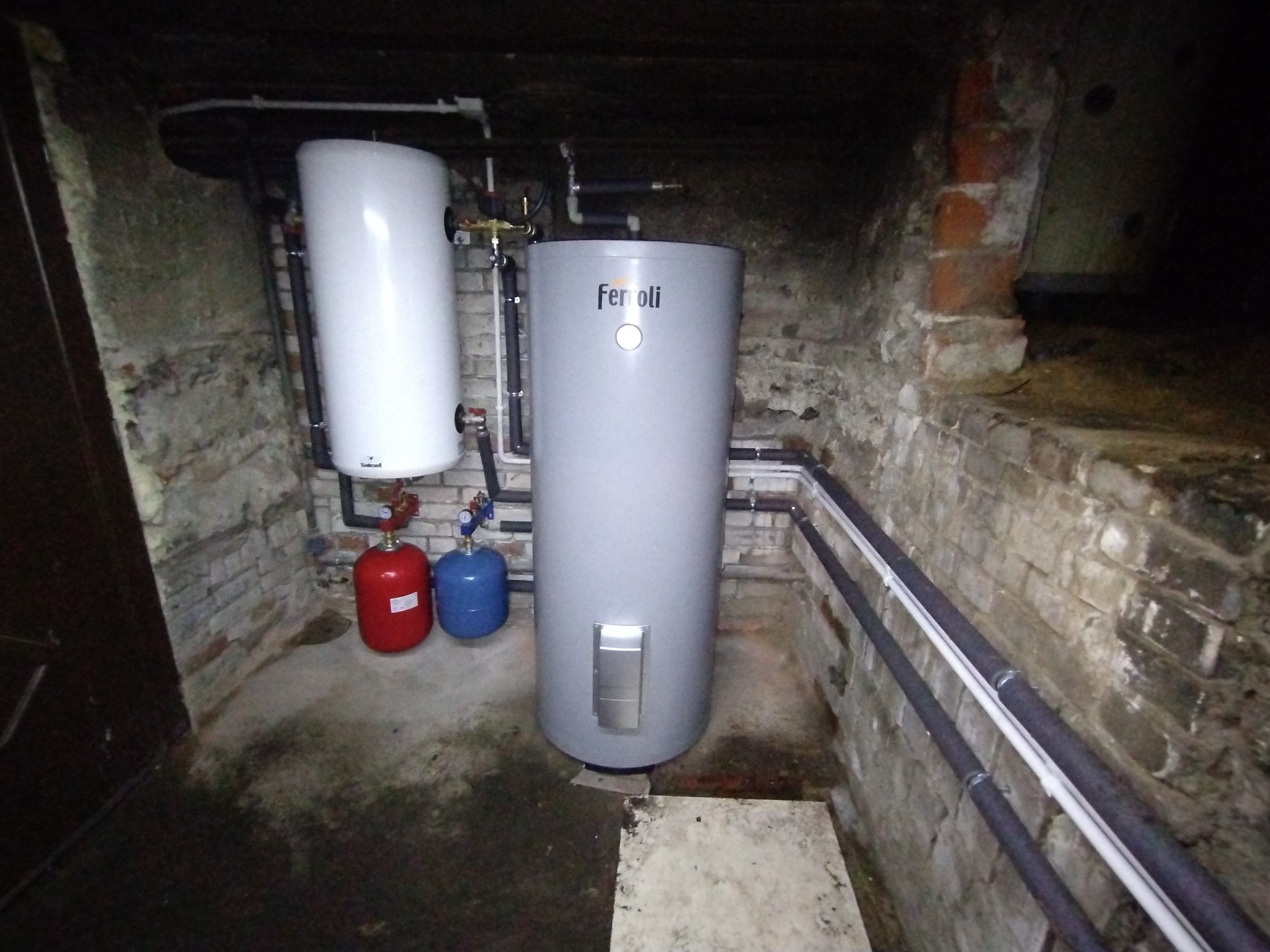 Dwa zbiorniki buforowe w piwnicy z ceglanymi ścianami, jeden biały, drugi szary z logo Ferroli, obok czerwony i niebieski zbiornik wyrównawczy.