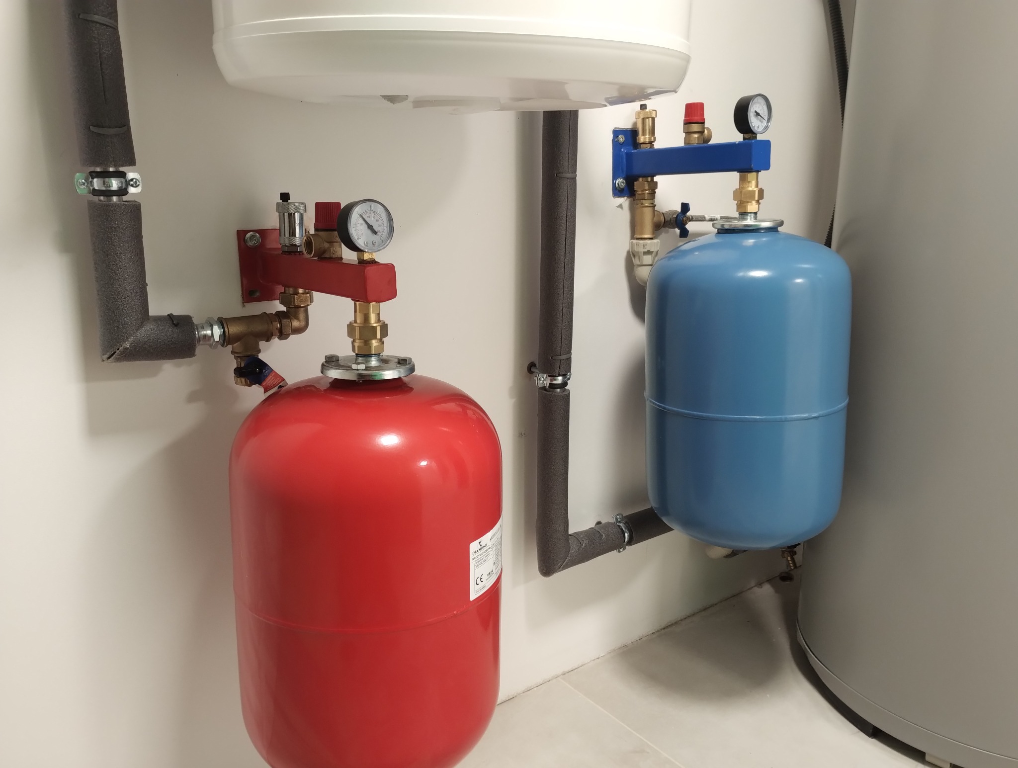 Czerwony i niebieski zbiornik ciśnieniowy z manometrami, zamontowane na ścianie, z widoczną izolacją rur.