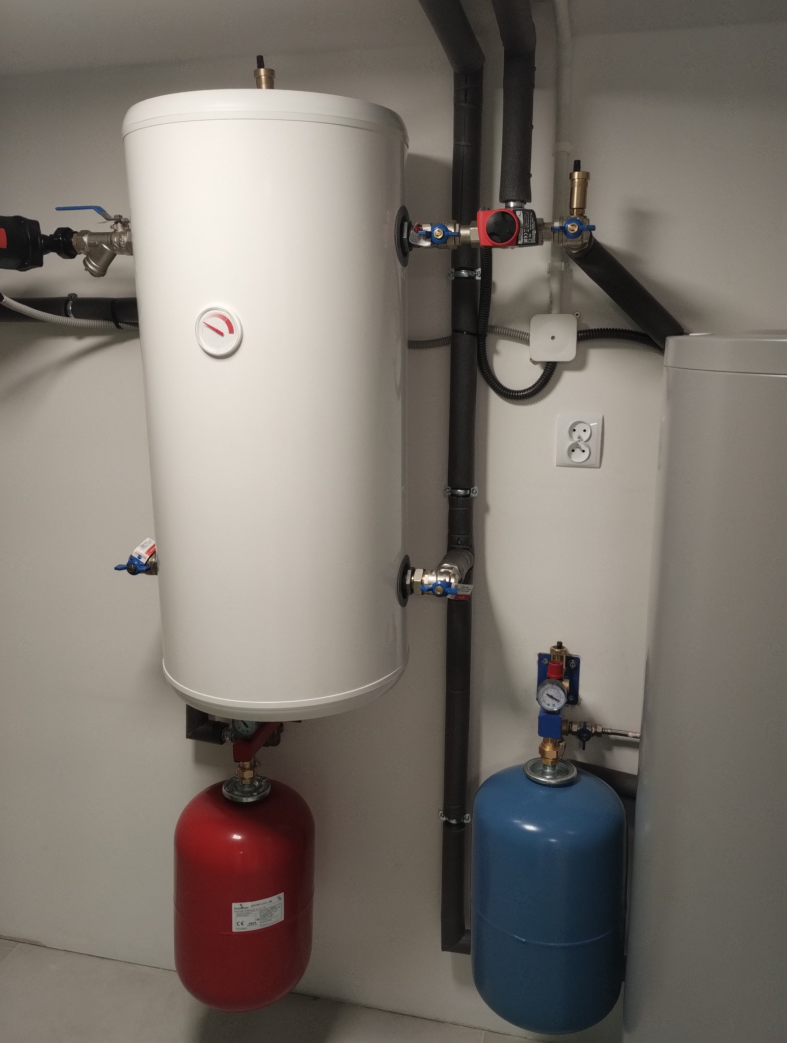 Biały zasobnik ciepłej wody z czerwonym manometrem, podłączony do czerwonego naczynia przeponowego i instalacji grzewczej z czarnymi rurami, obok niebieskie naczynie przeponowe z manometrem.