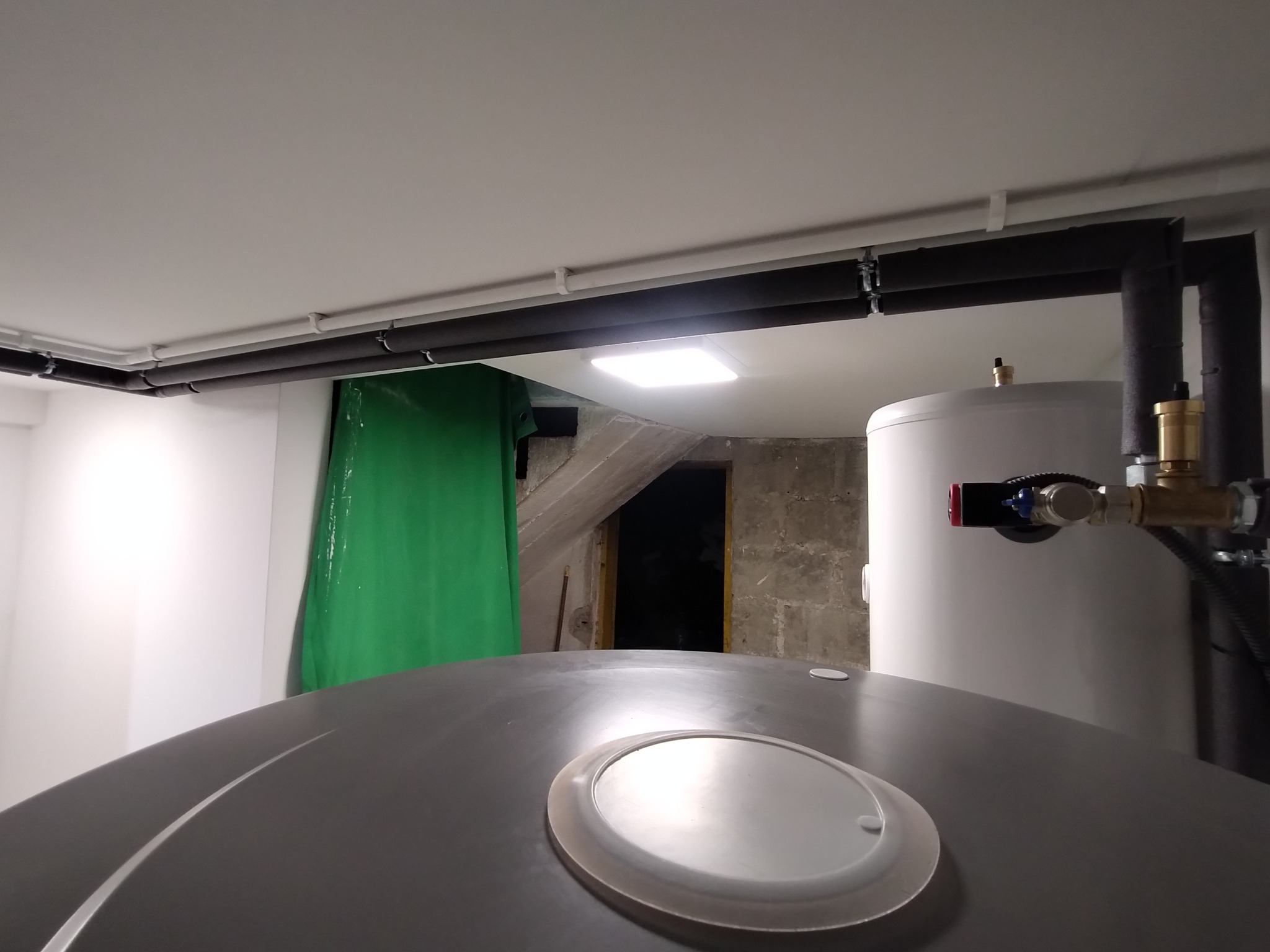 Dwa zbiorniki akumulacyjne w piwnicy, widoczne rury izolowane termicznie, zielona folia ochronna na ścianie, surowe ściany w tle.