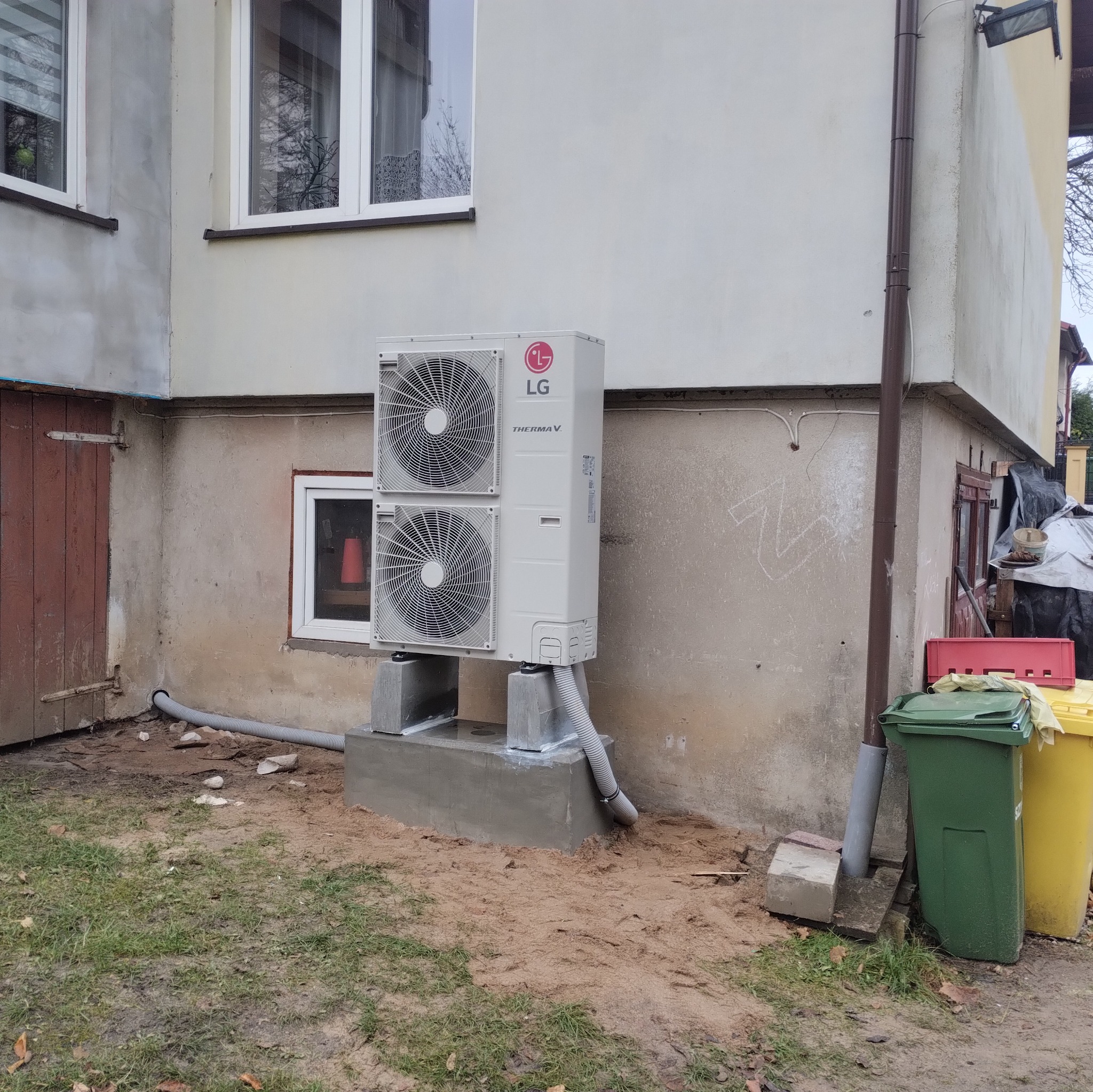 Zewnętrzna jednostka pompy ciepła LG Therma V, zamontowana na betonowych podstawach przy ścianie budynku z widocznymi rurami odprowadzającymi skropliny i elementami instalacji.