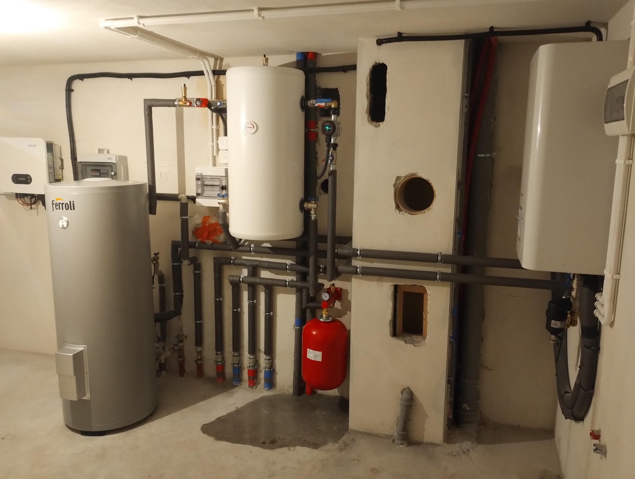 Kotłownia z szarym zbiornikiem Ferroli, białym zasobnikiem ciepłej wody, czerwonym naczyniem przeponowym i instalacją hydrauliczną w piwnicy, widoczne otwory w ścianie.