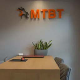 Recepcja biura z logo MTB na ścianie, biurkiem z laptopem i myszką oraz rośliną doniczkową.