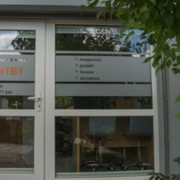 Wejście do biura rachunkowego MTBT z naklejonymi na szybie informacjami o świadczonych usługach: księgowość, podatki, finanse, doradztwo. W tle widoczne biurka i krzesła biurowe.