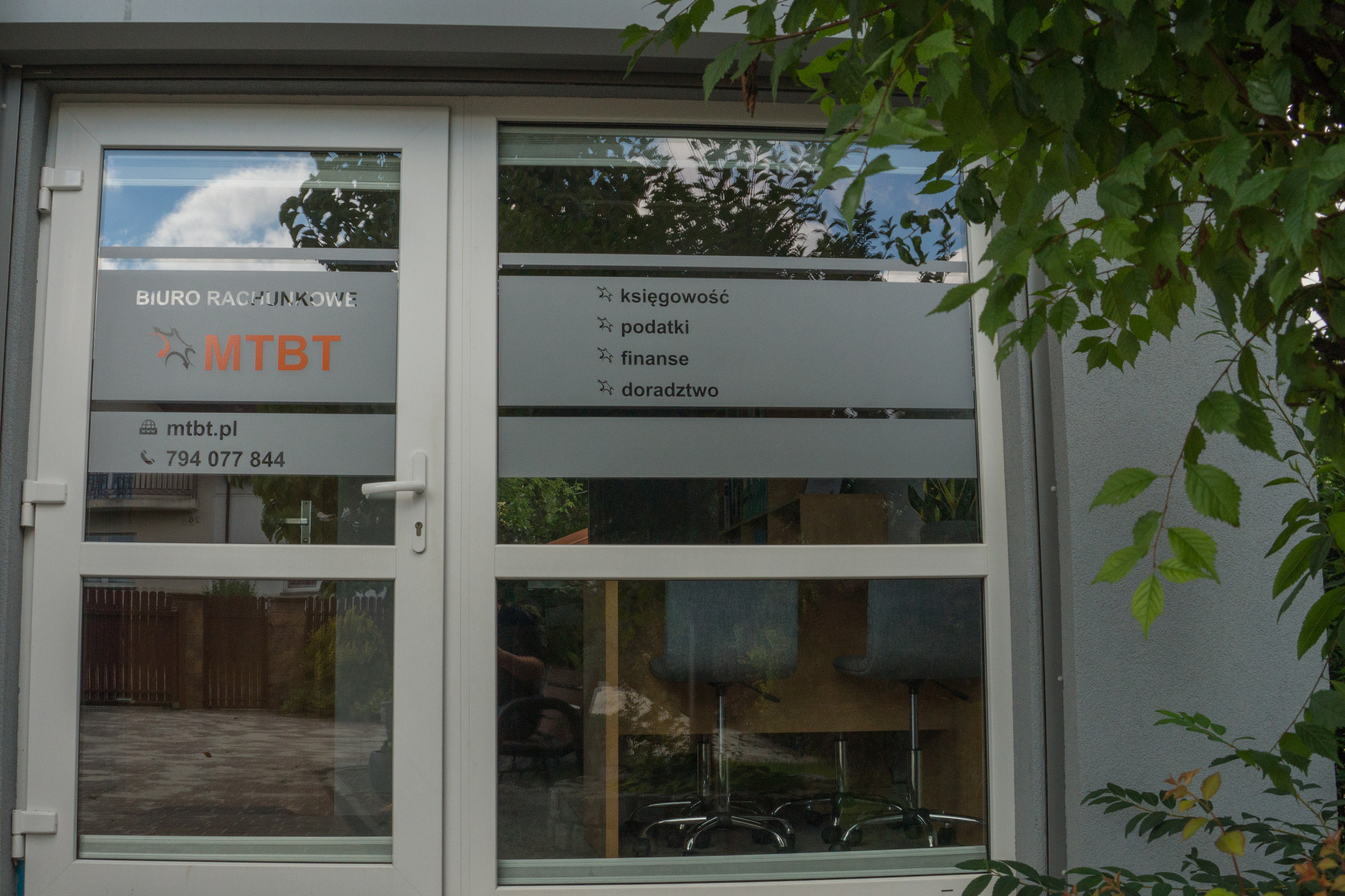 Wejście do biura rachunkowego MTBT z naklejonymi na szybie informacjami o świadczonych usługach: księgowość, podatki, finanse, doradztwo. W tle widoczne biurka i krzesła biurowe.