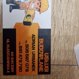 Wizytówka firmy budowlanej 'Usługi Budowlane Adrian Urbanek' z Żor, z kreskówkową grafiką budowlańca trzymającego poziomicę, leżąca na drewnianej powierzchni.