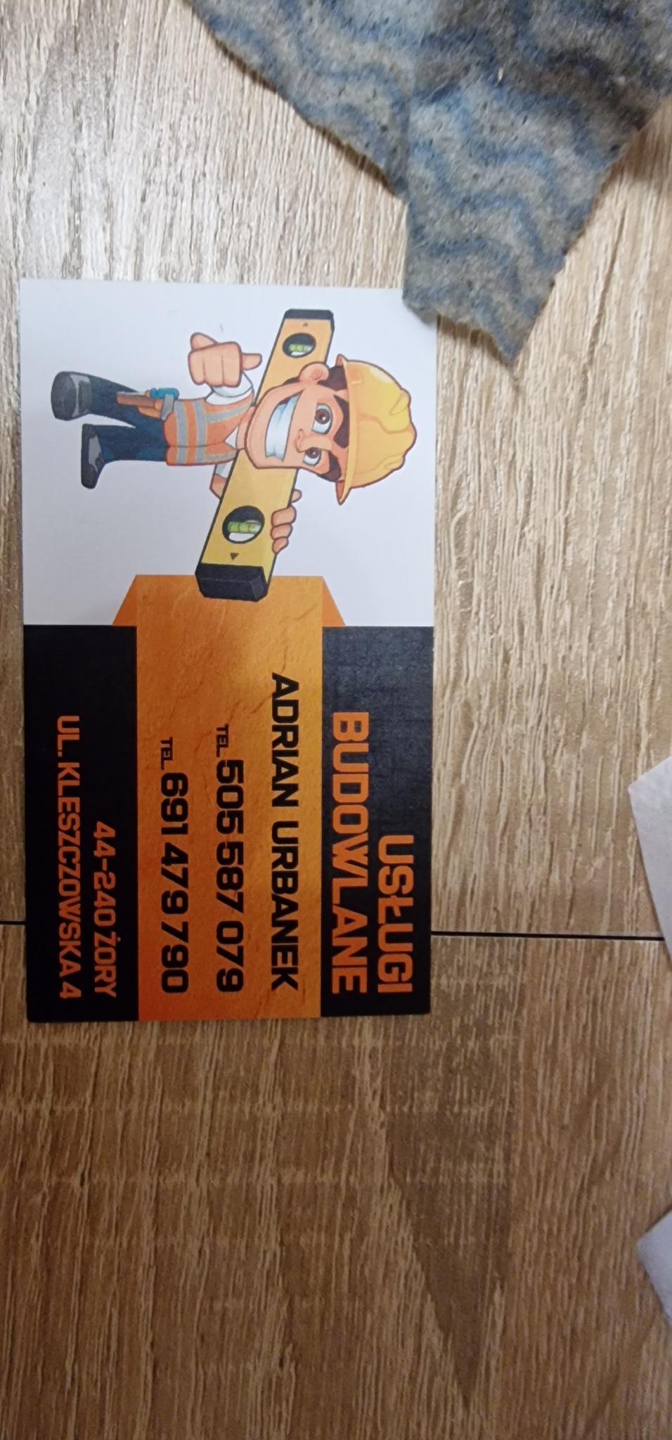 Wizytówka firmy budowlanej 'Usługi Budowlane Adrian Urbanek' z Żor, z kreskówkową grafiką budowlańca trzymającego poziomicę, leżąca na drewnianej powierzchni.
