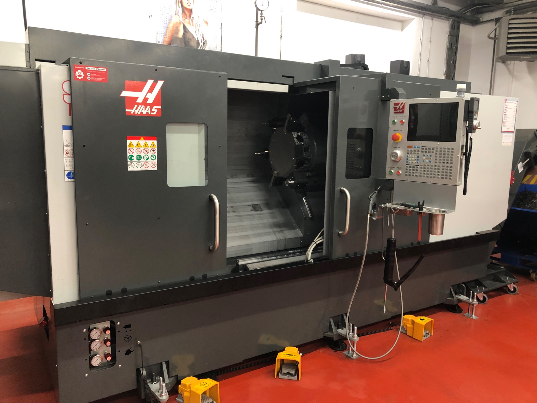 Szara, otwarta frezarka CNC marki HAAS z widocznym uchwytem narzędziowym wewnątrz, na czerwonej posadzce, z żółtymi pedałami sterującymi na podłodze.