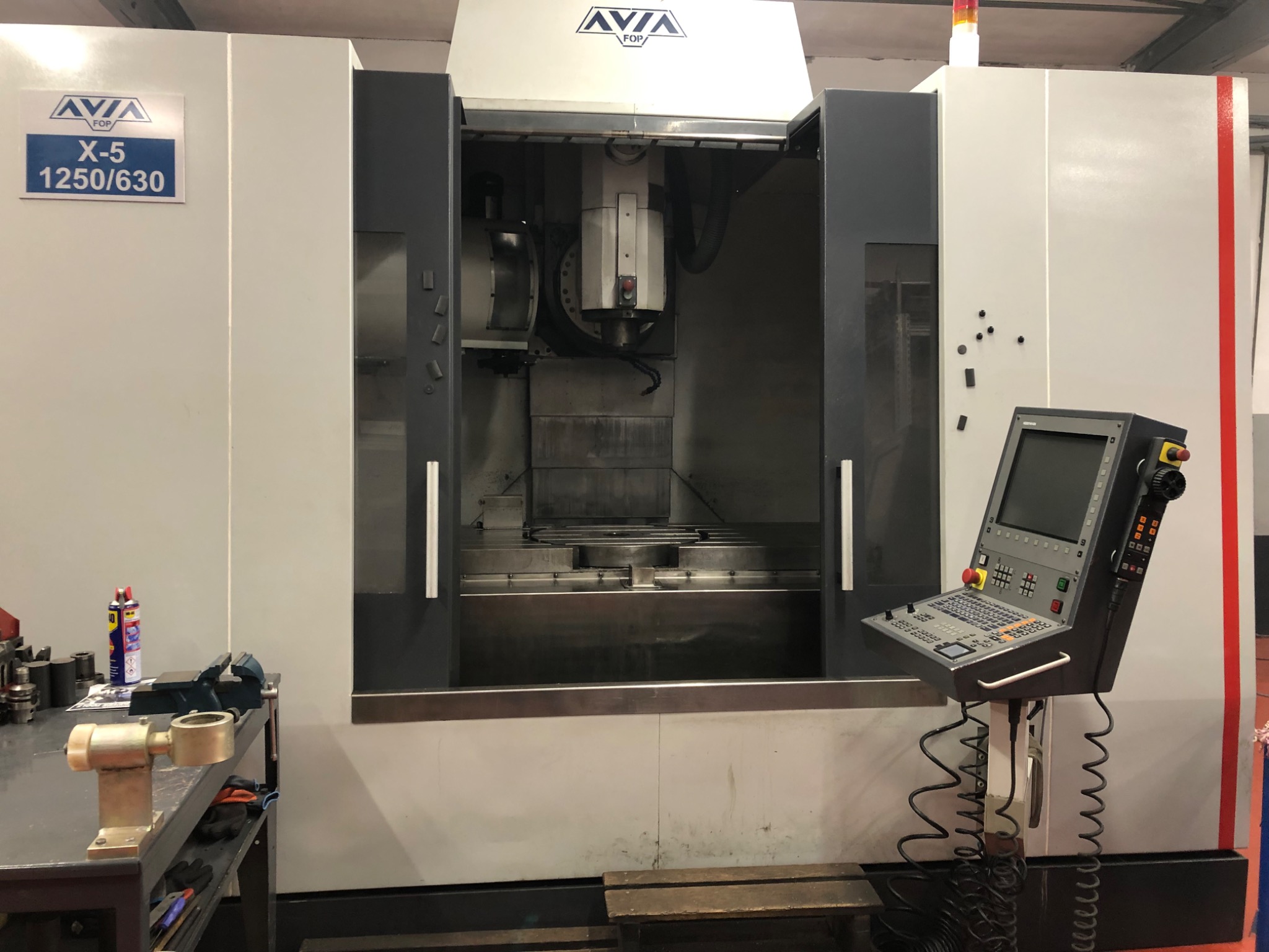 Duża frezarka CNC marki AVIA FOP X-5 1250/630 z otwartą komorą roboczą i panelem sterowania, widoczny stół roboczy z zamocowanymi elementami, obok na stole warsztatowym narzędzia i akcesoria.