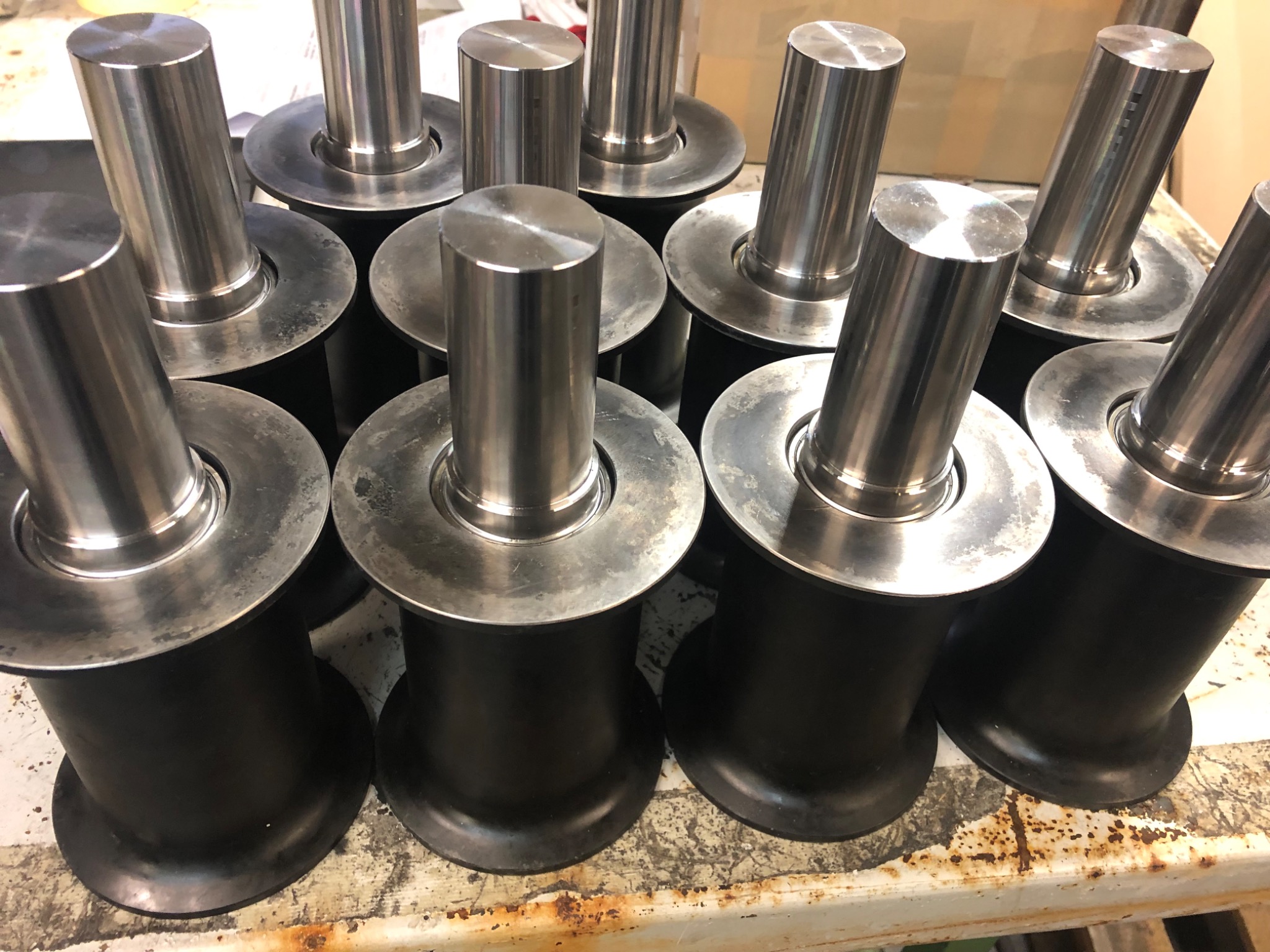 Grupowe ujęcie precyzyjnie obrobionych, metalowych elementów z kołnierzem, prawdopodobnie rolek do maszyn przemysłowych, ułożonych na zabrudzonym stole warsztatowym.