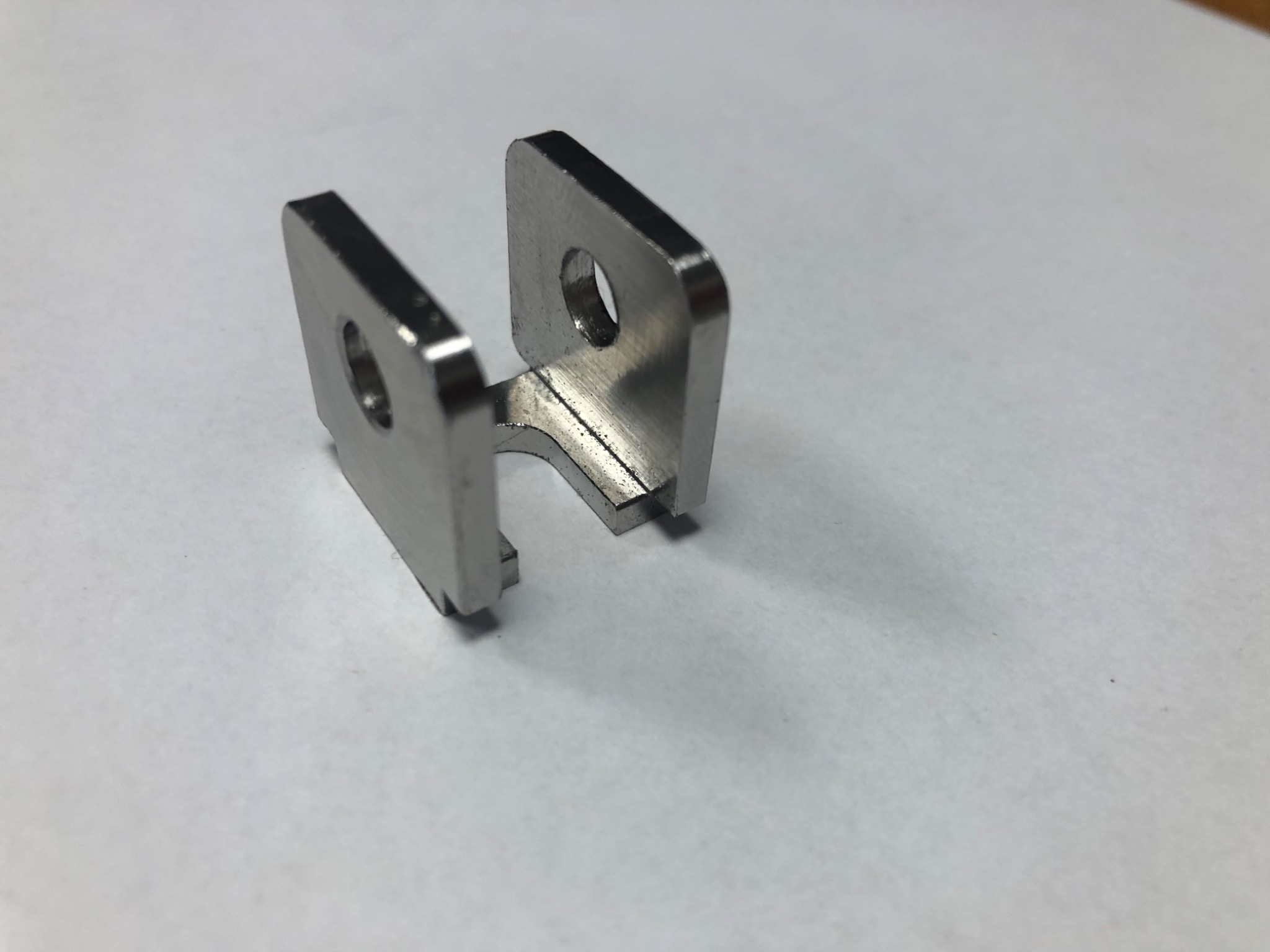 Metalowy element w kształcie litery U z otworami, wykonany z polerowanej stali, leżący na białym tle. Widoczne ślady obróbki skrawaniem.
