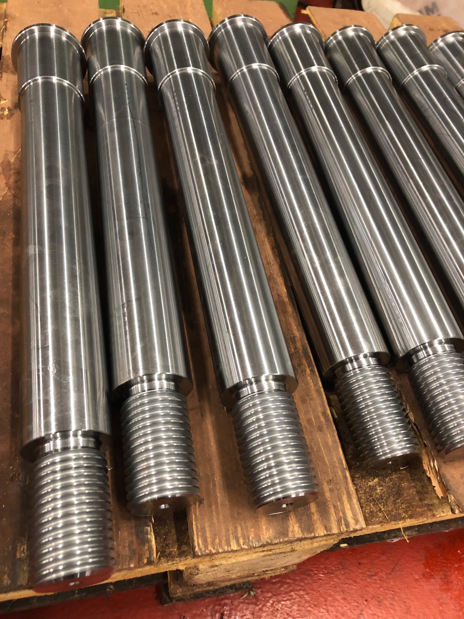 Rząd precyzyjnie obrobionych, gwintowanych stalowych wałów, ułożonych na drewnianej palecie, oświetlonych jasnym światłem.