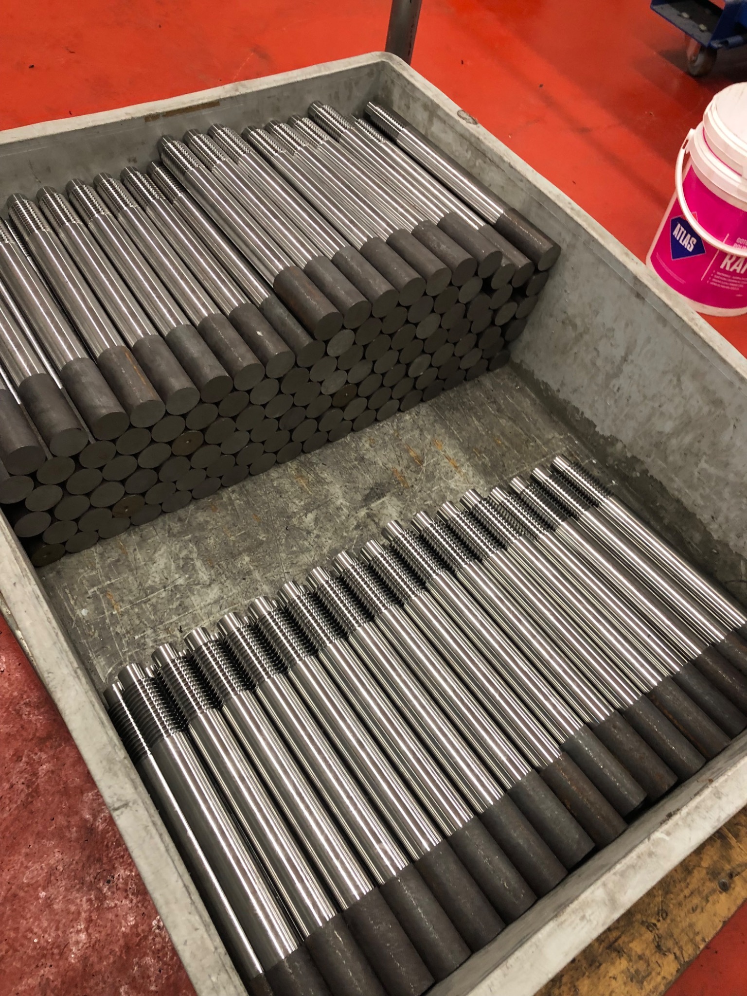 Partie obrobionych, gwintowanych prętów ze stali w szarym pojemniku w zakładzie w Grudziądzu. Część prętów ułożona równolegle, część składowana w stosie.