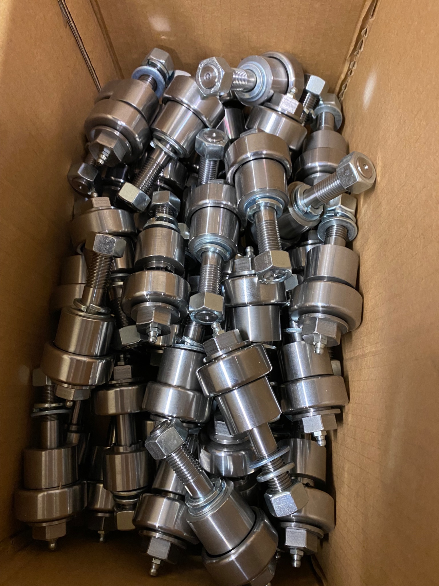 Partia srebrnych, metalowych rolek z gwintem i nakrętkami, ciasno upakowanych w kartonowym pudełku.