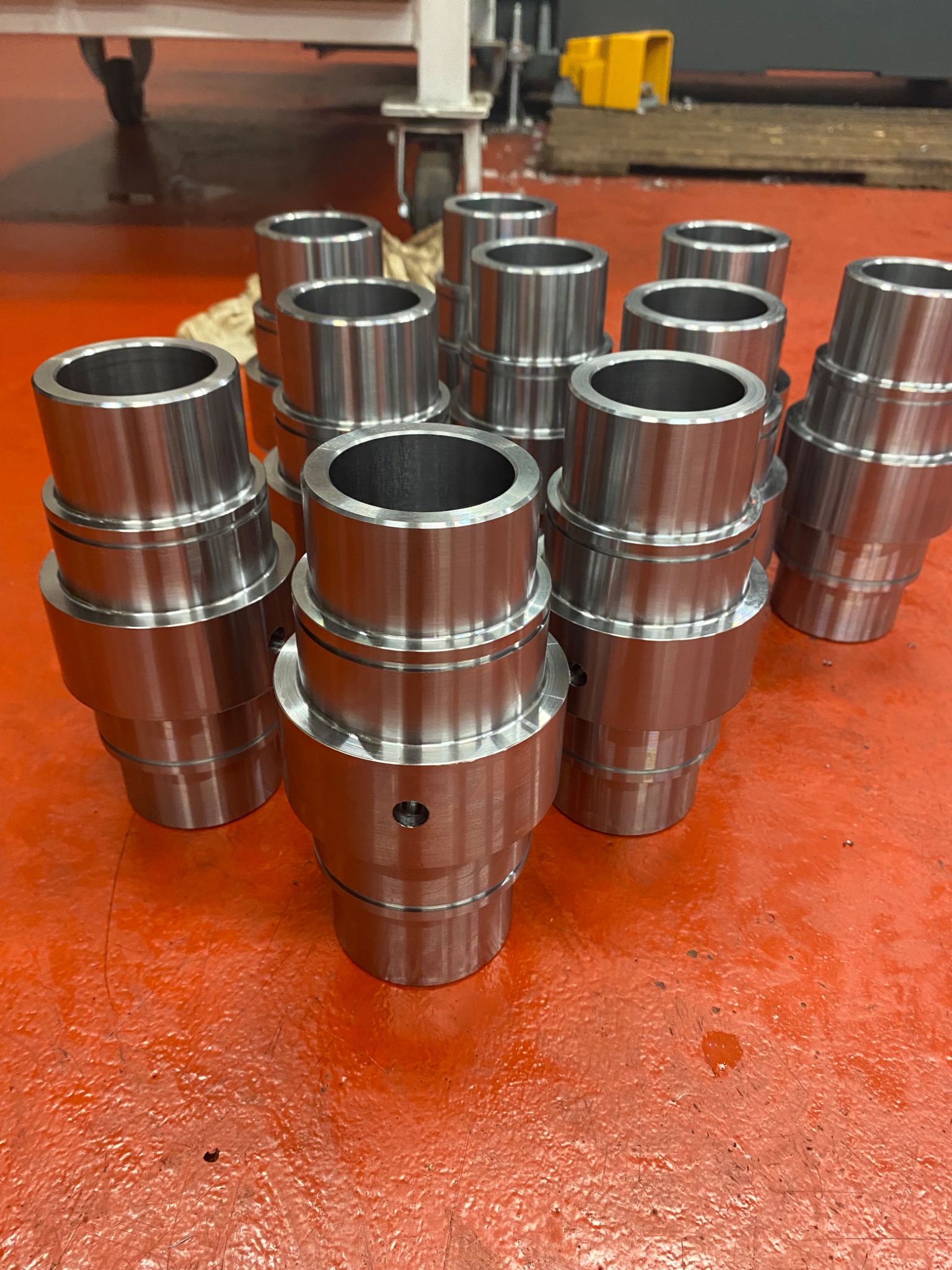 Partie obrobionych, metalowych tulei o cylindrycznym kształcie, ułożonych na pomarańczowej, przemysłowej podłodze. Widoczne ślady obróbki i otwory technologiczne.