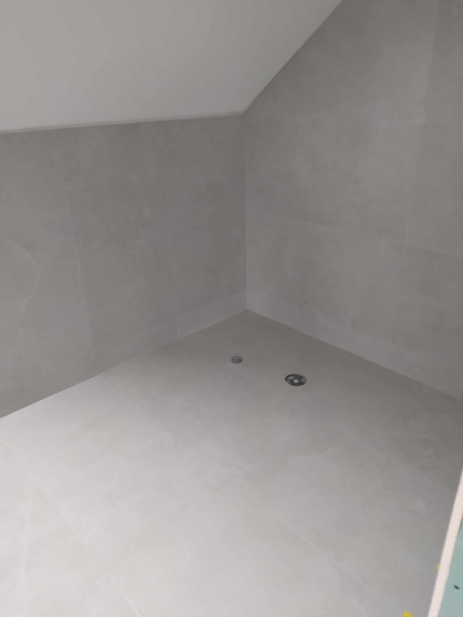 Wykończona kabina prysznicowa z szarymi płytkami na ścianach i białymi płytkami na podłodze, widoczne dwa odpływy liniowe.