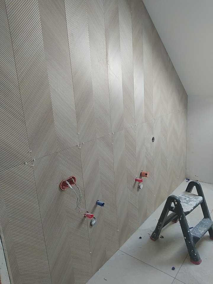 Ściana w trakcie wykańczania z wzorem chevron, widoczne wystające kable i rurki instalacyjne oraz drabinka malarska.