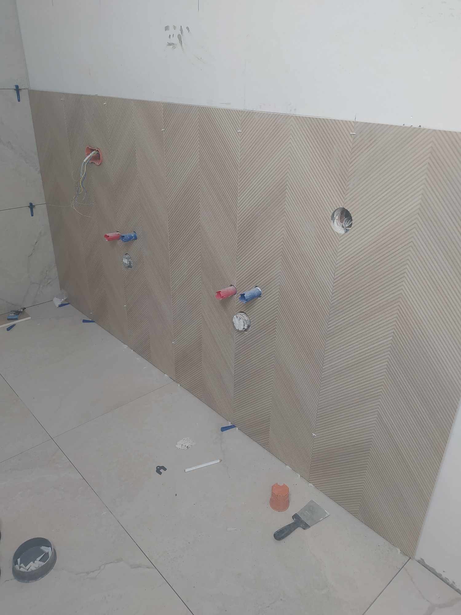Ściana z ułożonymi płytkami w jodełkę, widoczne otwory na instalacje wodne i elektryczne, oraz narzędzia glazurnicze na podłodze podczas prac wykończeniowych.