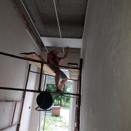 Valeria BUD Vasyl Trachuk - Pracownik na rusztowaniu montuje konstrukcję pod sufit podwieszany w nowym pomieszczeniu z widocznym oknem i elementami instalacji elektrycznej.