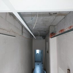 Valeria BUD Vasyl Trachuk - Korytarz w trakcie prac wykończeniowych: zamontowane profile sufitowe, ściany po wstępnym tynkowaniu, widoczna instalacja elektryczna, w głębi widoczna łazienka z białymi płytkami.