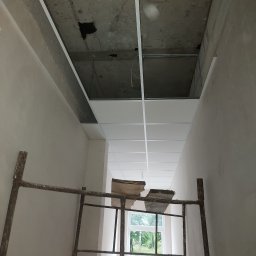 Valeria BUD Vasyl Trachuk - Montaż sufitu podwieszanego w trakcie prac, widoczne stelaże i częściowo ułożone płyty, rusztowanie w pomieszczeniu z oknem.