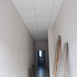 Valeria BUD Vasyl Trachuk - Korytarz w trakcie remontu: surowe ściany, podwieszany sufit kasetonowy, drewniane deski opierające się o ścianę, widok w głąb pomieszczenia.