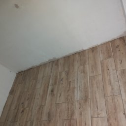 Valeria BUD Vasyl Trachuk - Ułożone panele podłogowe imitujące drewno na ścianie w trakcie remontu, widoczne niedoskonałości przy suficie i ścianie, sufit biały, widać otwór po lampie.