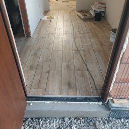 Valeria BUD Vasyl Trachuk - Ułożona podłoga z płytek imitujących drewno w nowym pomieszczeniu, widoczne worki z zaprawą, narzędzia i fragment wylewki z gruzem.