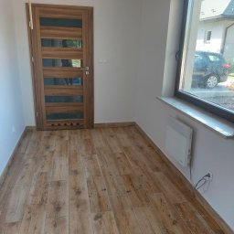 Valeria BUD Vasyl Trachuk - Wnętrze z drewnopodobnymi panelami podłogowymi, drewnianymi drzwiami z przeszkleniami i grzejnikiem elektrycznym pod oknem.