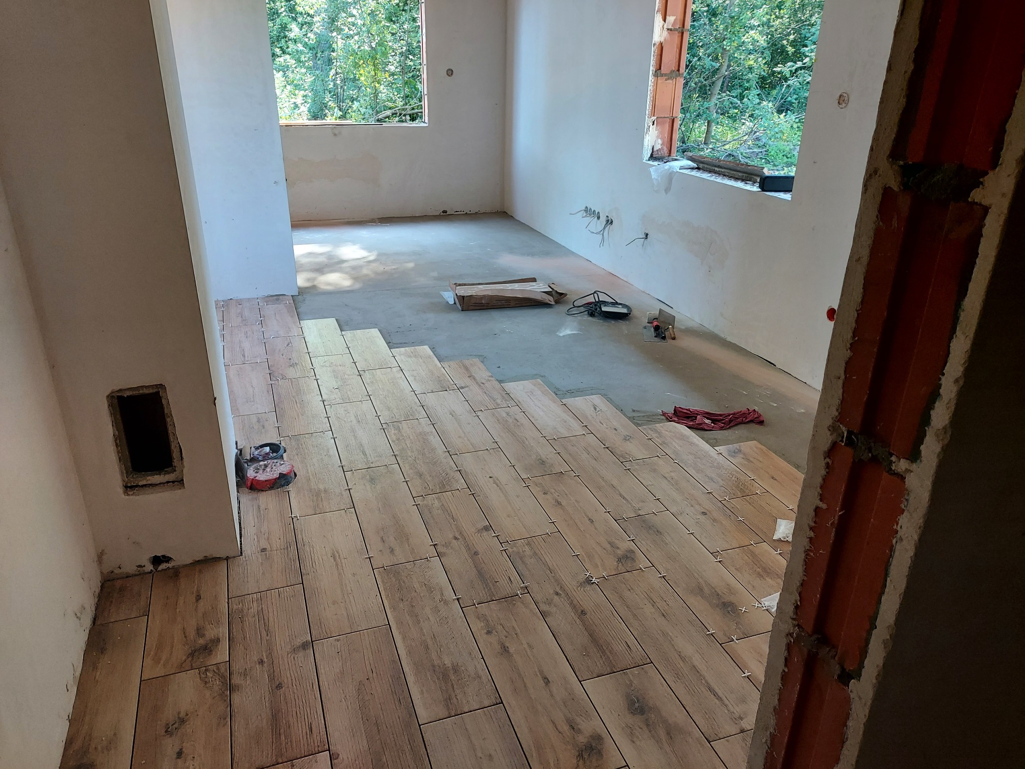 Układanie płytek podłogowych imitujących drewno w jasnym pomieszczeniu z dwoma oknami, widoczne narzędzia i akcesoria glazurnicze, etap prac wykończeniowych.