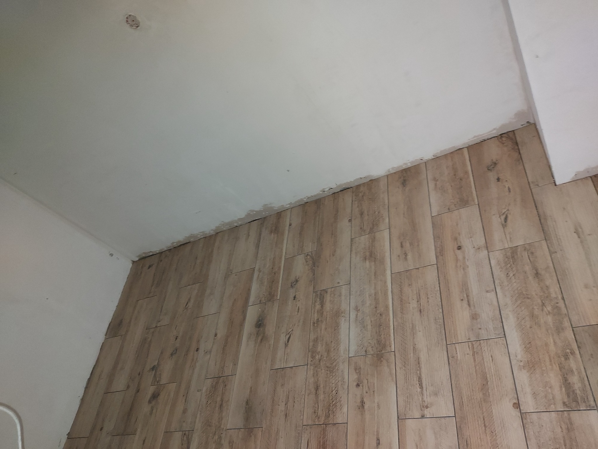 Ułożone panele podłogowe imitujące drewno na ścianie w trakcie remontu, widoczne niedoskonałości przy suficie i ścianie, sufit biały, widać otwór po lampie.