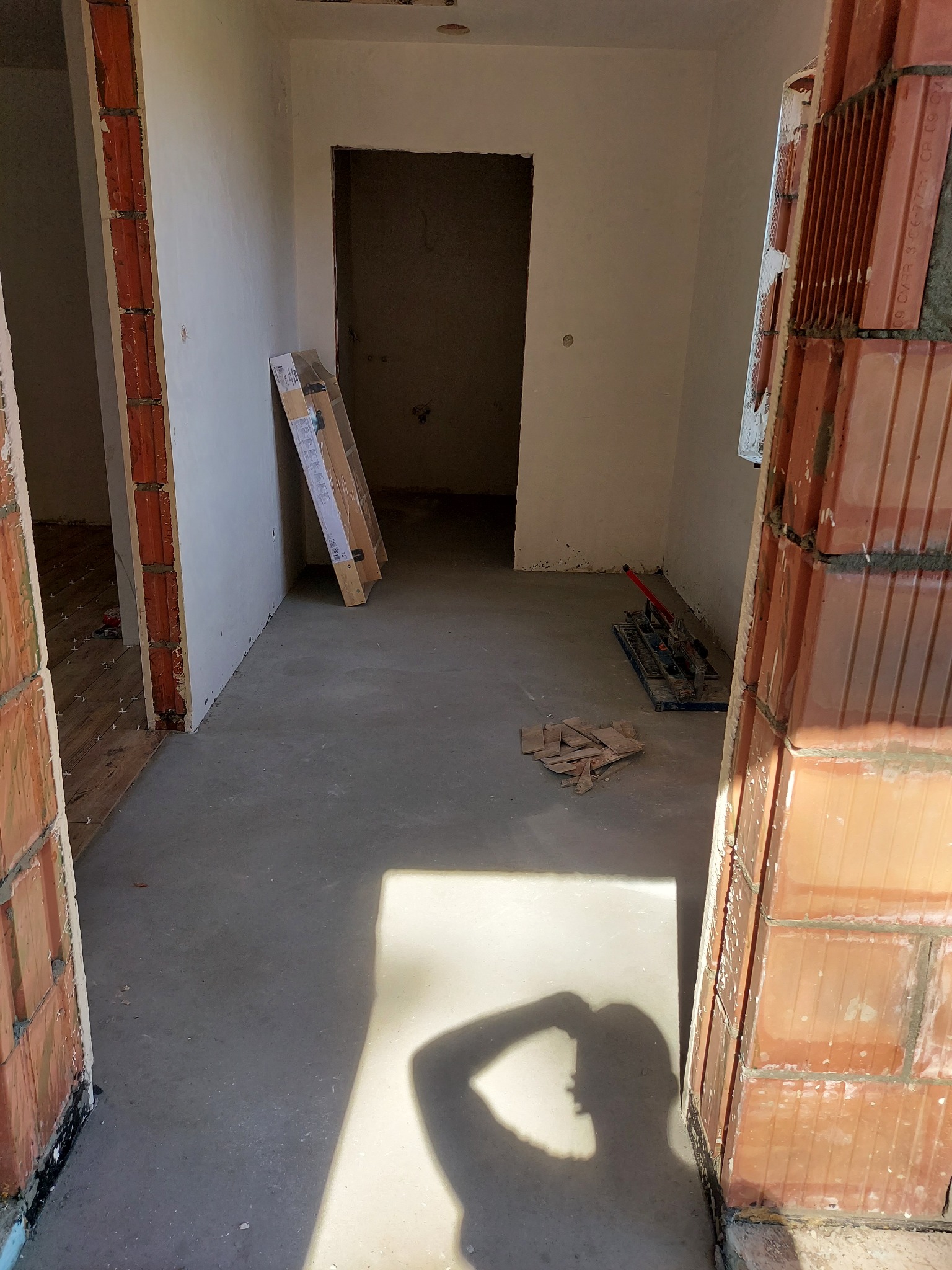 Wnętrze w trakcie prac budowlanych: surowe ściany, wylewka podłogowa, widoczne bloczki ceramiczne na ościeżnicach i narzędzia do cięcia płytek w tle, cień osoby fotografującej na podłodze.
