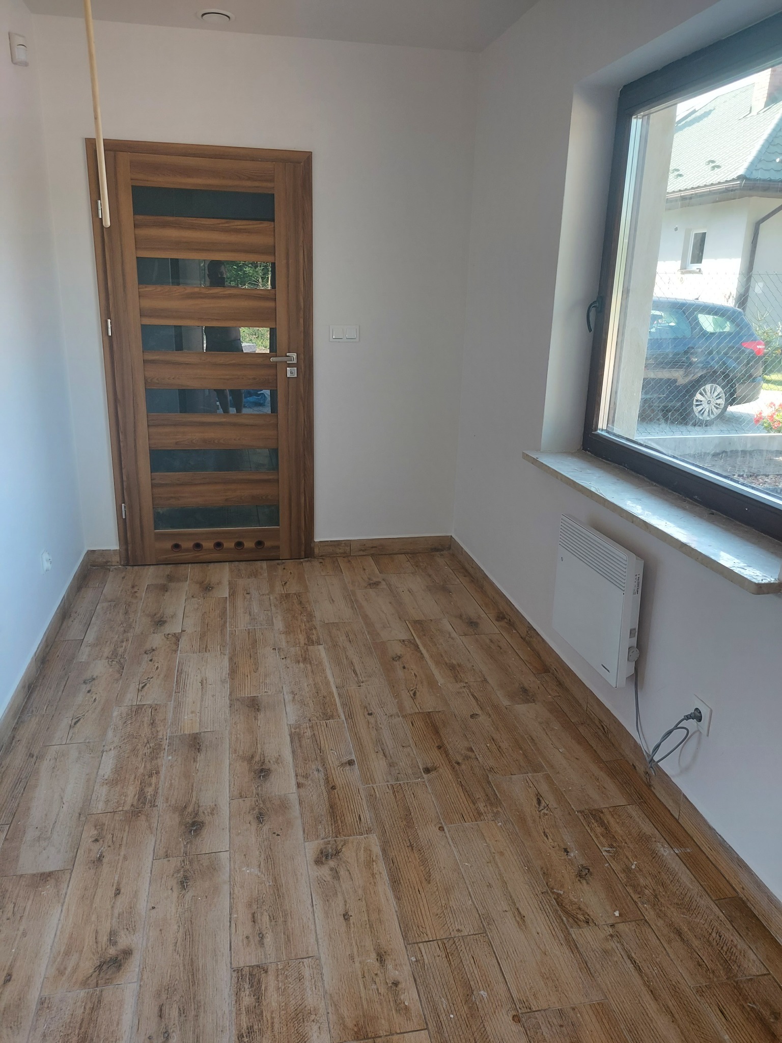 Wnętrze z drewnopodobnymi panelami podłogowymi, drewnianymi drzwiami z przeszkleniami i grzejnikiem elektrycznym pod oknem.