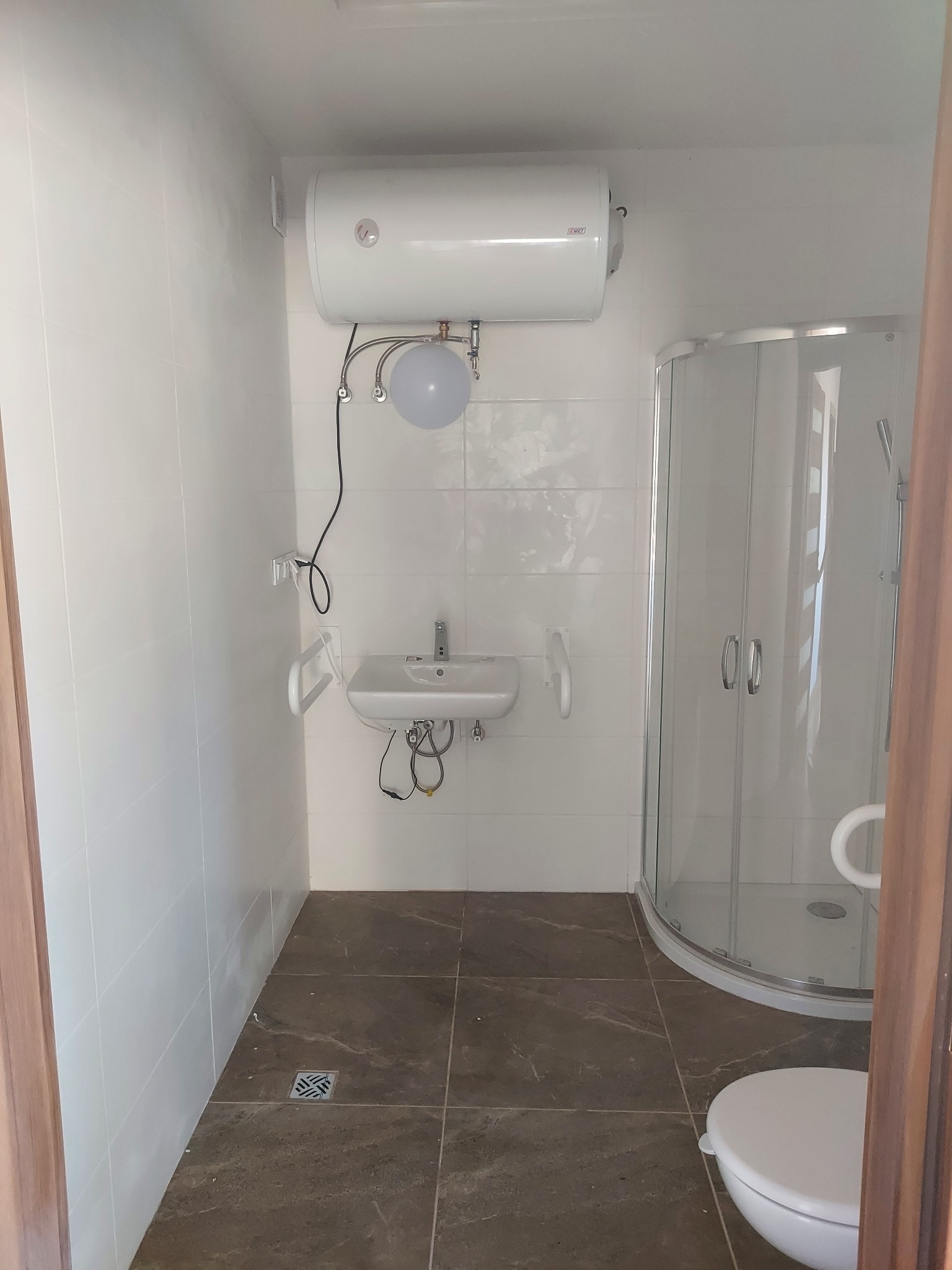 Wykończona łazienka z białymi płytkami, brązową podłogą, kabiną prysznicową, umywalką, bojlerem i uchwytami dla osób niepełnosprawnych.