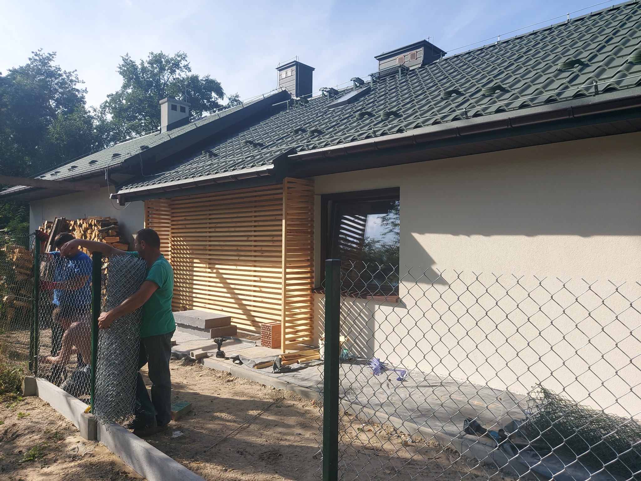 Montaż ogrodzenia z siatki na posesji z drewnianą elewacją i ułożonym drewnem na boku, widoczne narzędzia i materiały budowlane.