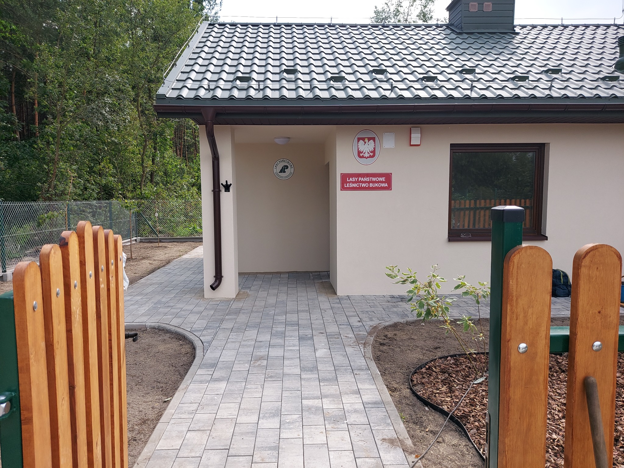 Nowo wybudowany budynek Lasów Państwowych Leśnictwo Bukowa z szarym dachem, beżową elewacją i utwardzonym terenem z kostki brukowej, widok zza drewnianego ogrodzenia.