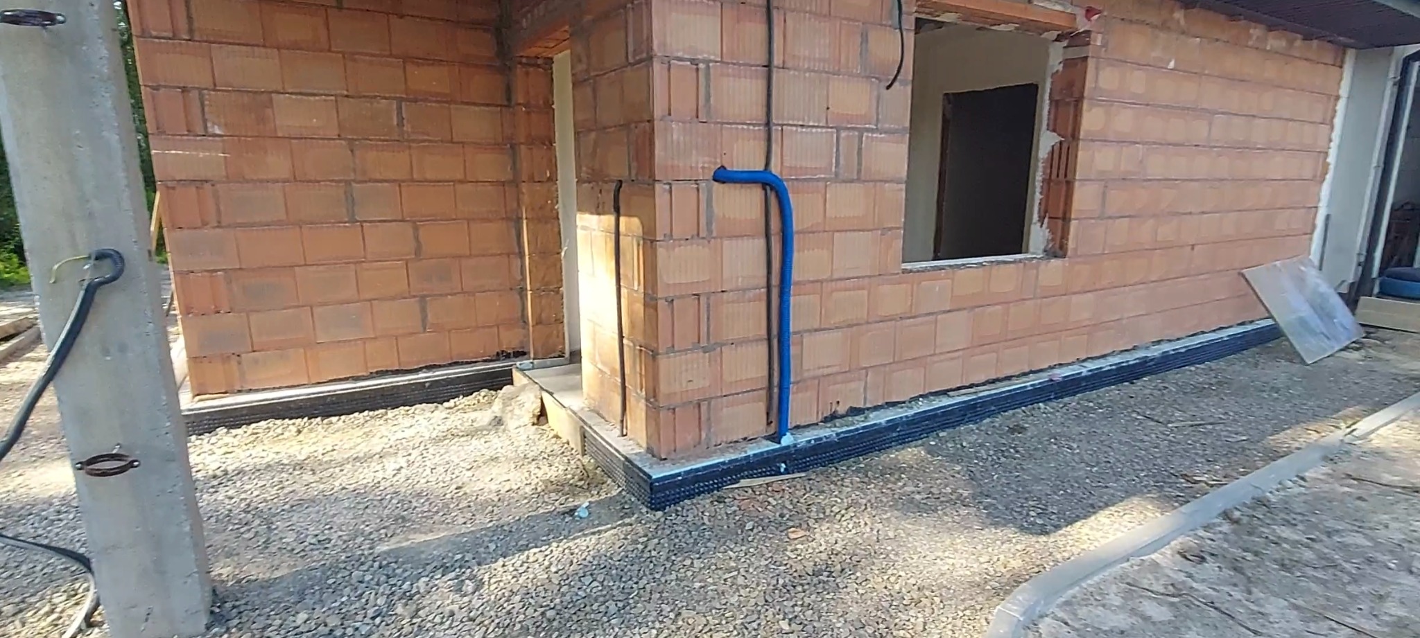 Budowa domu: ściany z czerwonej cegły ceramicznej Porotherm, widoczna niebieska rura instalacyjna na zewnątrz budynku, fundament z izolacją przeciwwilgociową.