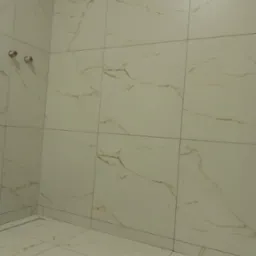 Wykończona łazienka z jasnymi, marmurkowymi płytkami na ścianach i podłodze, widoczne przyłącza wodne na ścianie i odpływ liniowy w podłodze.