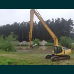 Żółta koparka nabiera urobek z płytkiego stawu, w tle widoczne pryzmy ziemi i las.