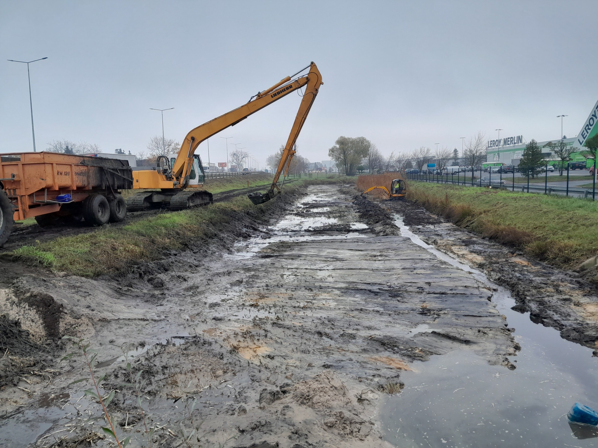 Żółta koparka Liebherr i pomarańczowa przyczepa na brzegu wykopanego, błotnistego rowu, w tle widoczny budynek Leroy Merlin, pochmurny dzień.