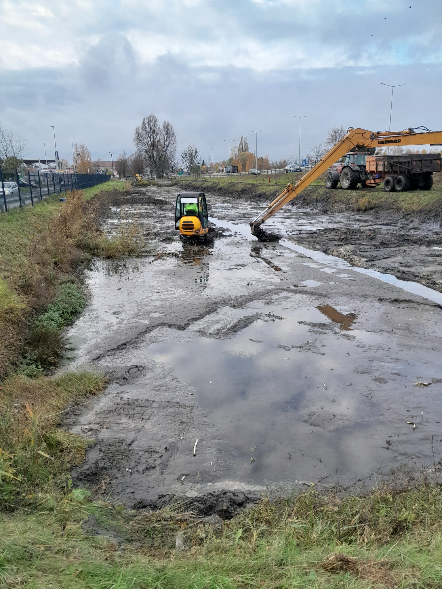 Żółta minikoparka Liebherr w błotnistym wykopie, z odbiciami chmur w wodzie, obok stoi traktor z przyczepą, w tle widoczna droga i zabudowania.