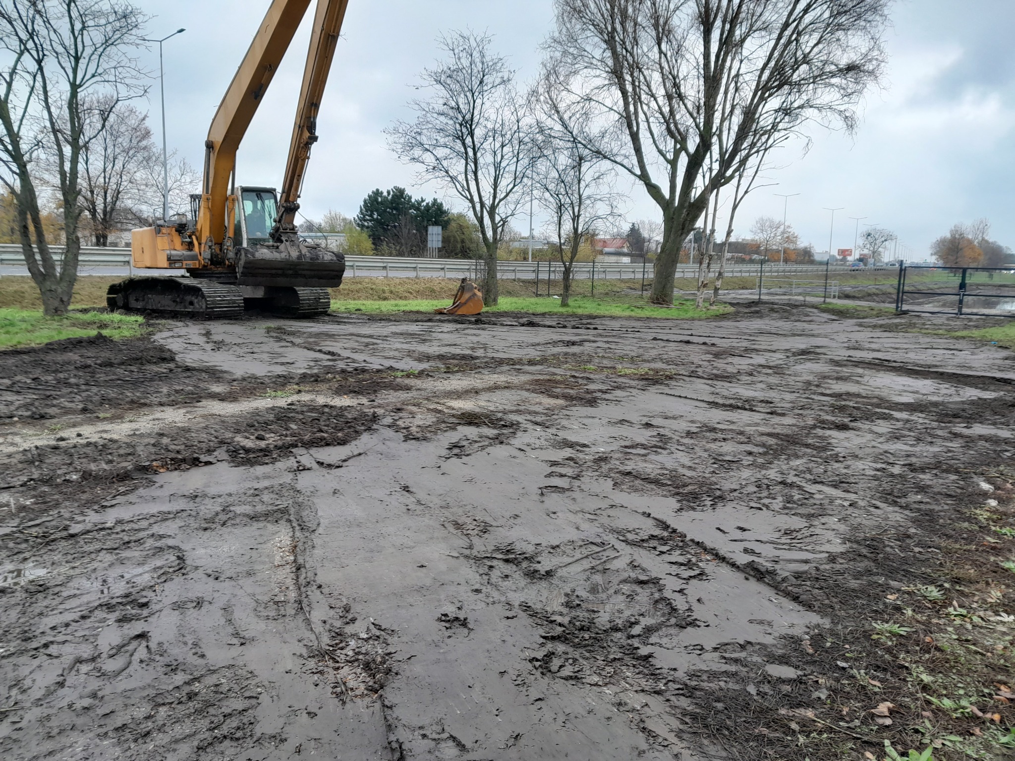 Żółta koparka gąsienicowa na błotnistym terenie, widoczne ślady opon, w tle drzewa bez liści, bariery drogowe i pochmurne niebo.