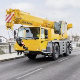 Żółty dźwig samojezdny Liebherr LTM 1040-2.1 z wysięgnikiem skierowanym w górę, stojący na asfaltowej drodze na tle zabudowań miejskich i pochmurnego nieba.