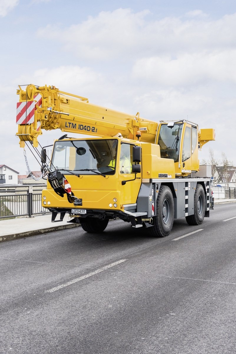 Żółty dźwig samojezdny Liebherr LTM 1040-2.1 z wysięgnikiem skierowanym w górę, stojący na asfaltowej drodze na tle zabudowań miejskich i pochmurnego nieba.