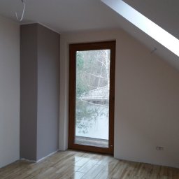 PAW-REMONT - Pomalowane na kremowo ściany poddasza z oknem w drewnianej ramie, fragment pomalowanej na szaro kolumny i błyszcząca podłoga imitująca drewno.