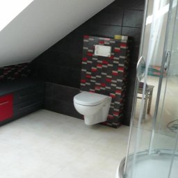 PAW-REMONT - Wykończona łazienka na poddaszu z toaletą podwieszaną na ścianie wyłożonej mozaiką w odcieniach szarości i czerwieni, obok kabina prysznicowa z przezroczystego szkła.