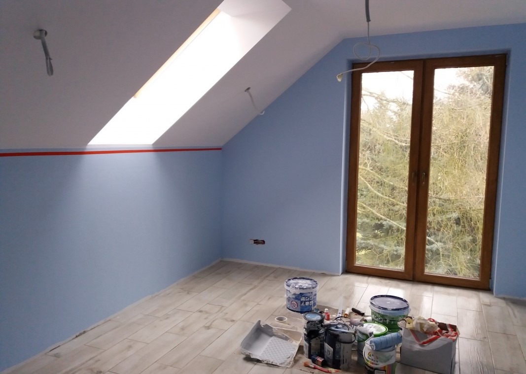 Poddasze ze skośnym oknem dachowym, pomalowane na jasny niebieski kolor, z widocznymi puszkami farb i narzędziami malarskimi na podłodze z paneli imitujących drewno, zabezpieczona czerwona linia...
