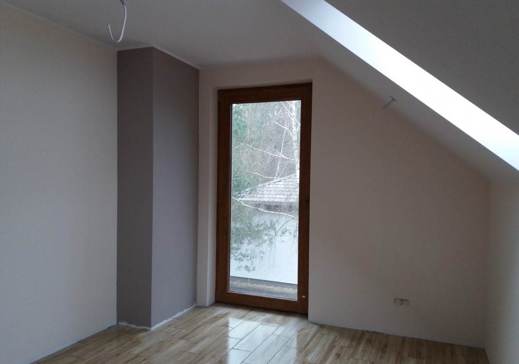 Pomalowane na kremowo ściany poddasza z oknem w drewnianej ramie, fragment pomalowanej na szaro kolumny i błyszcząca podłoga imitująca drewno.