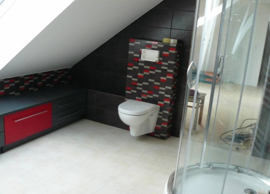 Wykończona łazienka na poddaszu z toaletą podwieszaną na ścianie wyłożonej mozaiką w odcieniach szarości i czerwieni, obok kabina prysznicowa z przezroczystego szkła.