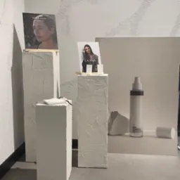 Minimalistyczna kompozycja z trzema białymi postumentami o chropowatej fakturze, na których umieszczono zdjęcia portretowe i kosmetyki La'Ana oraz Team D'Joseph, z białym tłem i neutralną kolorystyką.